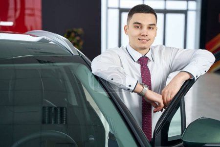 Smiling man stands leaning against open car door, workplace manager in car elephant