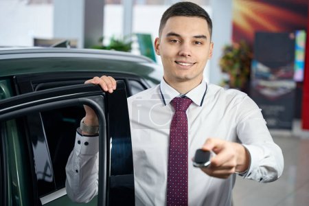 Téléchargez les photos : Charmant jeune homme debout près d'une voiture ouverte avec des clés de voiture, gestionnaire en vêtements décontractés - en image libre de droit