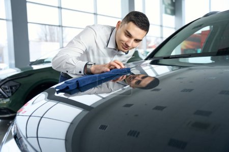 Photo for Smiling workplace manager in car showroom wiping car hood, view windows indoors - Royalty Free Image