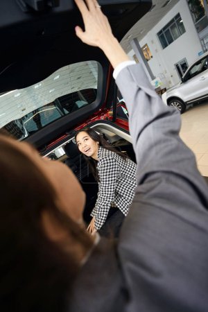 Téléchargez les photos : Sourire femme d'affaires intéressée dans la veste de poule assise dans le coffre de la voiture lève les yeux - en image libre de droit
