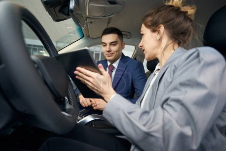 Foto de Hombre y mujer en ropa de negocios sentados en asientos delanteros en automóvil y mirando en la tableta - Imagen libre de derechos