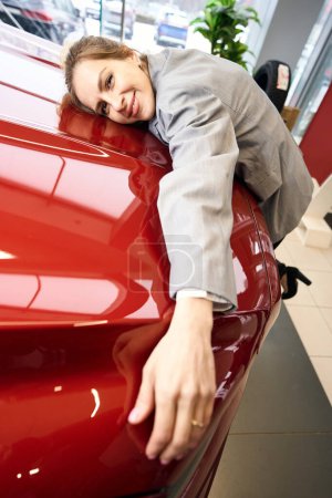 Lächelnde Dame umarmt Motorhaube und legt sich auf Auto im Autohaus