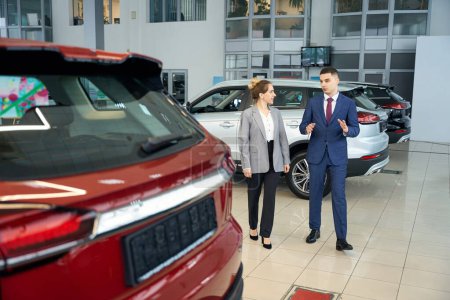 Foto de Feliz hombre de negocios hablando con la mujer de traje y de pie cerca de coche rojo en la concesionaria - Imagen libre de derechos