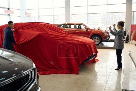 Photo for Happy lady holding hands up and man in suit and standing near automobile under fabric in car dealership - Royalty Free Image