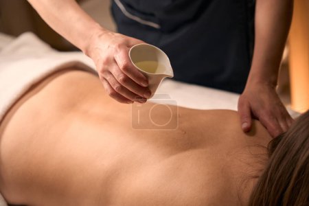 Photo for Cropped photo of skilled masseuse pouring organic oil from ceramic jug on naked back of adult woman - Royalty Free Image
