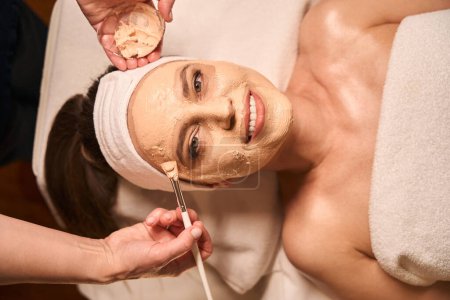 Téléchargez les photos : Vue du dessus de la cliente souriante lors de l'application du masque facial réalisée par un professionnel des soins de la peau - en image libre de droit