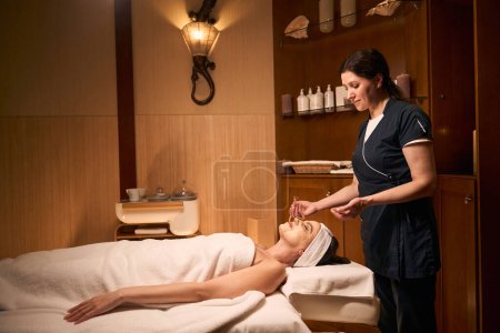 Photo for Focused beautician with brush in hand standing over calm female client lying on couch during mask application - Royalty Free Image