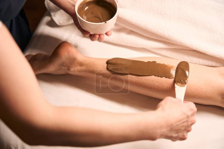 Photo for Cropped photo of esthetician applying seaweed paste to woman lower leg with spatula - Royalty Free Image
