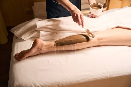 Foto de Foto recortada de cosmético aplicando pasta de algas marrones a la pierna femenina spa con espátula - Imagen libre de derechos