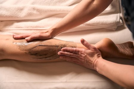 Foto de Foto recortada de manos masoterapeutas frotando la pierna inferior del cliente femenino con pasta de algas marrones - Imagen libre de derechos
