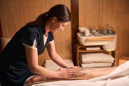 Foto de Masoterapeuta enfocado frotando pasta de algas marrones en la piel en la pierna inferior de la mujer - Imagen libre de derechos