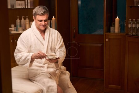 Téléchargez les photos : Homme calme en peignoir assis sur le canapé avec tasse de tisane dans le centre de bien-être - en image libre de droit