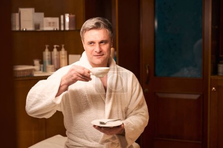 Photo for Waist-up portrait of tranquil spa client in bathrobe seated on couch lifting cup of drink to his lips - Royalty Free Image