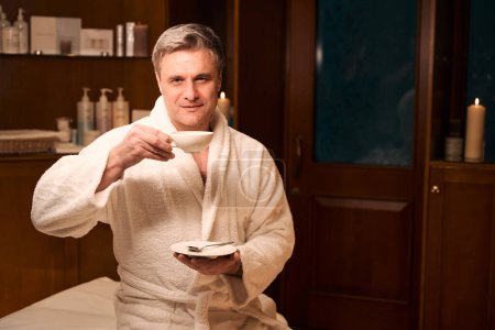 Photo for Serene mature man dressed in bathrobe seated on massage table lifting cup of beverage to mouth - Royalty Free Image