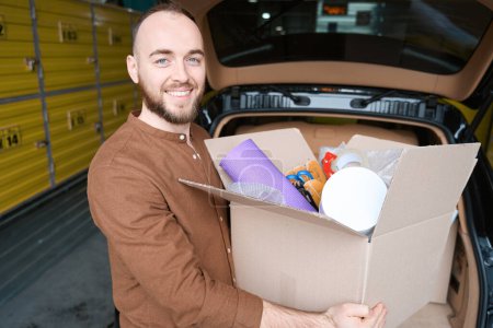 Téléchargez les photos : Homme souriant debout près de la voiture dans l'entrepôt avec une boîte dans ses mains - en image libre de droit