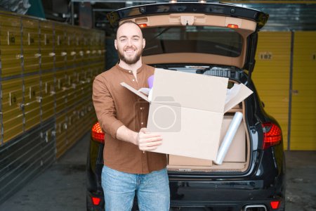 Photo for Satisfied caucasian man holding a package, standing near the car in the storage room - Royalty Free Image