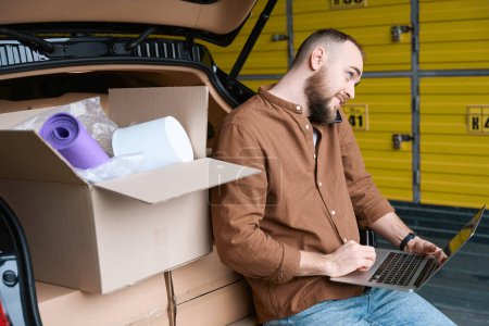 Téléchargez les photos : Homme satisfait adulte avec un ordinateur portable parlant au téléphone près d'une voiture dans un entrepôt - en image libre de droit