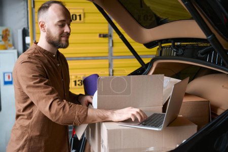 Téléchargez les photos : L'homme attentif regarde un ordinateur portable, debout près du coffre d'une voiture avec des marchandises dans un entrepôt - en image libre de droit