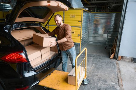 Téléchargez les photos : Satisfait jeune homme chargeant des boîtes en carton dans le coffre d'une voiture dans un entrepôt - en image libre de droit