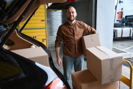 Photo for Adorable grown man standing near the trunk of a car in a warehouse with a box in his hands - Royalty Free Image