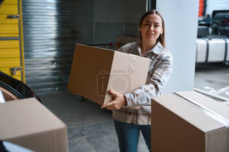 Photo for Smiling young female person standing in a warehouse holding a box in her hands - Royalty Free Image