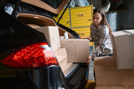 Téléchargez les photos : Femme concentrée chargeant des marchandises dans des boîtes dans le coffre d'une voiture alors qu'elle se tenait dans un entrepôt - en image libre de droit