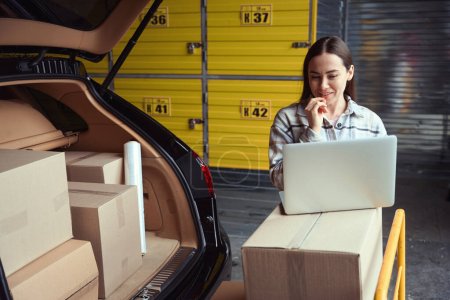 Téléchargez les photos : Femme adulte souriante regardant un ordinateur portable debout dans l'entrepôt près du coffre des boîtes - en image libre de droit