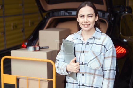 Photo for Nice woman holding a laptop while standing in a warehouse near a truck with a load - Royalty Free Image