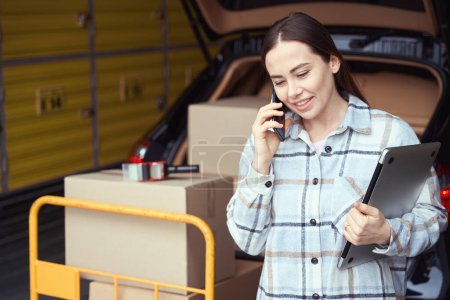 Téléchargez les photos : Joyeuse dame ayant une conservation au téléphone tout en se tenant dans l'entrepôt près de la voiture avec des boîtes - en image libre de droit