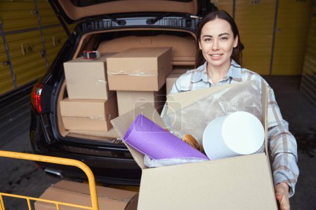 Téléchargez les photos : Femme agréable restant derrière le coffre de la voiture dans l'entrepôt et tenant une boîte en carton avec des choses - en image libre de droit