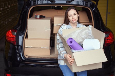 Téléchargez les photos : Jeune femme occupée debout près du coffre d'une voiture et gardant la boîte avec des trucs dans un entrepôt - en image libre de droit