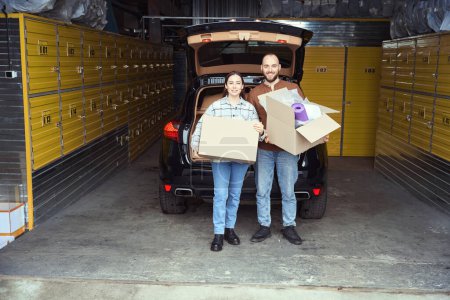 Téléchargez les photos : Heureux homme et femme debout près de la voiture dans un entrepôt et tenant des boîtes de choses dans leurs mains - en image libre de droit