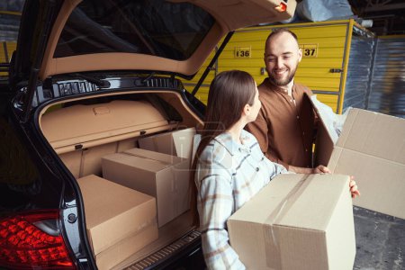 Photo for Happy man and woman looking at each other, standing with boxes in their hands near a car in a warehouse - Royalty Free Image