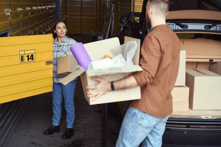 Téléchargez les photos : Jeune homme et femme debout dans un entrepôt près d'une voiture avec des boîtes dans leurs mains - en image libre de droit