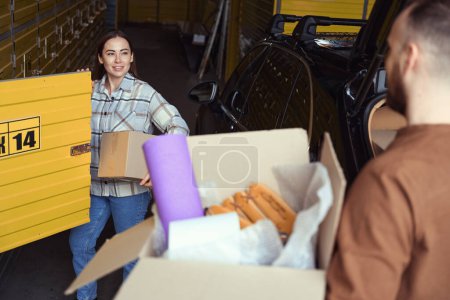 Photo for Two busy people standing with boxes in their hands near a car in a warehouse - Royalty Free Image