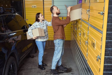Téléchargez les photos : Deux personnes occupées empilant une boîte de choses dans un casier de stockage tout en se tenant près d'une voiture - en image libre de droit