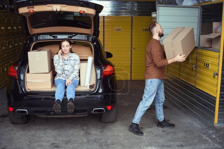 Photo for Grown female sitting in the car trunk and speaking on mobile phone while busy man putting box in the storage - Royalty Free Image