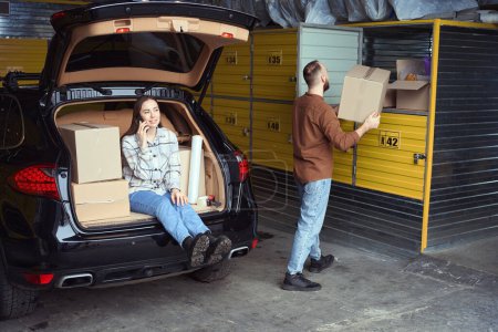 Photo for Satisfied woman sitting in the car trunk and speaking on mobile phone while adult man stacking merchandise for storage - Royalty Free Image