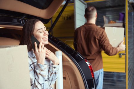Téléchargez les photos : Jeune femme souriante parlant sur un smartphone dans le coffre de la voiture à côté d'un homme avec une boîte dans les mains - en image libre de droit