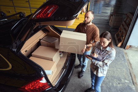 Photo for Focused man holding a box near the trunk of a car while a woman writing something down - Royalty Free Image