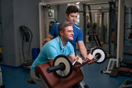 Photo for Focused fit male performing seated barbell curl on weight bench under guidance of fitness coach - Royalty Free Image