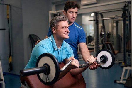 Photo for Smiling mature man doing barbell curl on weight bench guided by gym instructor - Royalty Free Image