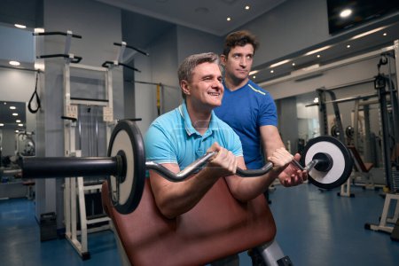 Téléchargez les photos : Joyeuse forme masculine effectuant la boucle d'haltère sur le banc de poids sous le contrôle de l'entraîneur de forme physique - en image libre de droit
