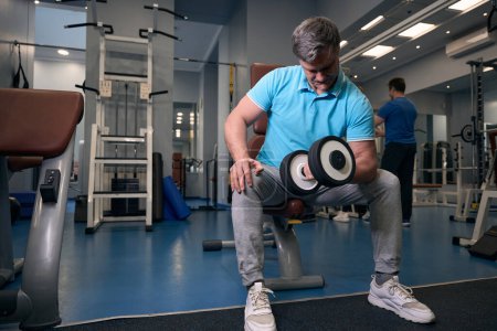 Téléchargez les photos : Homme adulte musclé effectuant une boucle d'haltère assis à un bras pendant l'entraînement de gymnastique - en image libre de droit