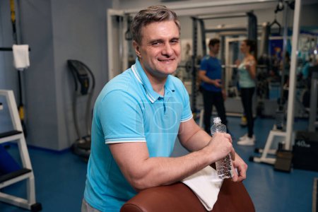 Téléchargez les photos : Homme athlétique heureux avec une bouteille d'eau ouverte appuyée sur un banc d'exercice au gymnase - en image libre de droit