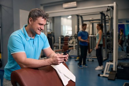 Photo for Smiling athletic man with mobile phone in hands leaning on exercise bench in fitness club - Royalty Free Image