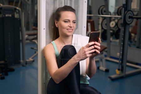 Téléchargez les photos : Agréable dame en forme lisant quelque chose sur l'écran tactile du smartphone tout en étant assis dans le club de fitness - en image libre de droit
