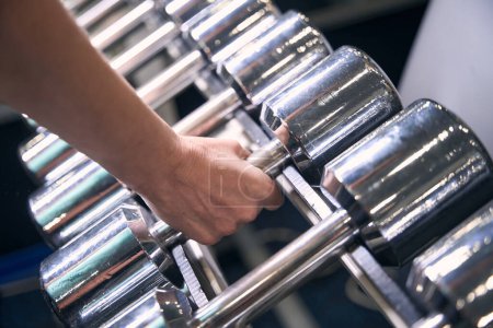 Photo for Cropped photo of bodybuilder hand picking up chrome-plated dumbbell from weight storage rack at gym - Royalty Free Image