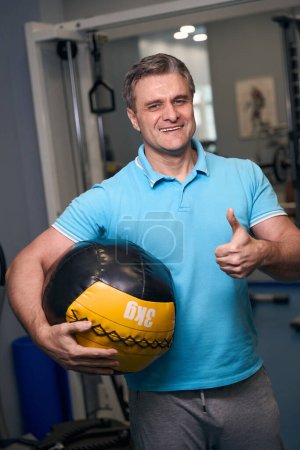 Foto de Hombre atlético sonriente con bola de goma debajo del brazo mostrando un gesto de pulgar hacia arriba ante la cámara en el gimnasio - Imagen libre de derechos