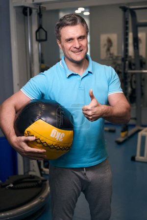 Foto de Alegre hombre deportivo sosteniendo la pelota de goma y haciendo pulgares hacia arriba signo delante de la cámara - Imagen libre de derechos